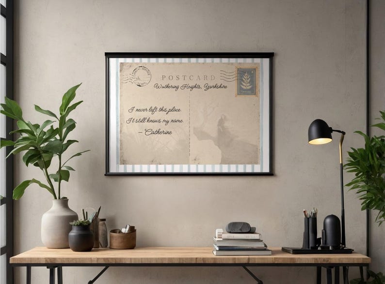May include: Framed vintage postcard print featuring the quote "I never left this place. It still knows my name. - Catherine." The postcard is beige with a black frame and a stamp design. The print is displayed on a wooden desk.