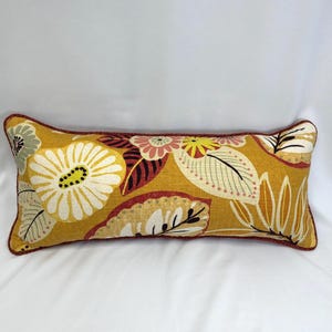 May include: Mustard yellow rectangular decorative pillow with a floral pattern. The pillow features large white flowers with yellow centers, and various leaves in shades of yellow, brown, and green. The edges are trimmed with a dark red piping.