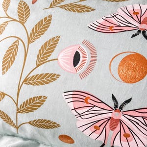 May include: A pale blue pillowcase featuring a repeating pattern of pink moths, flowers, and gold-coloured leaves. The design includes a stylized sun and small brown ovals. The pillowcase is made of a soft fabric.
