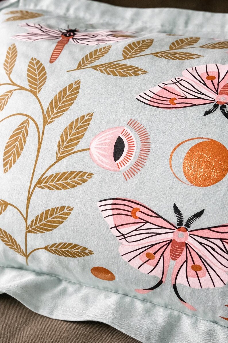 May include: A pale blue pillowcase featuring a repeating pattern of pink moths, flowers, and gold-coloured leaves. The design includes a stylized sun and small brown ovals. The pillowcase is made of a soft fabric.