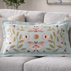 May include: Rectangular cushion with a light blue background and embroidered design. The design features moths, dragonflies, and floral elements in pink, gold, and brown. The cushion has a light blue border and is placed on a grey sofa.