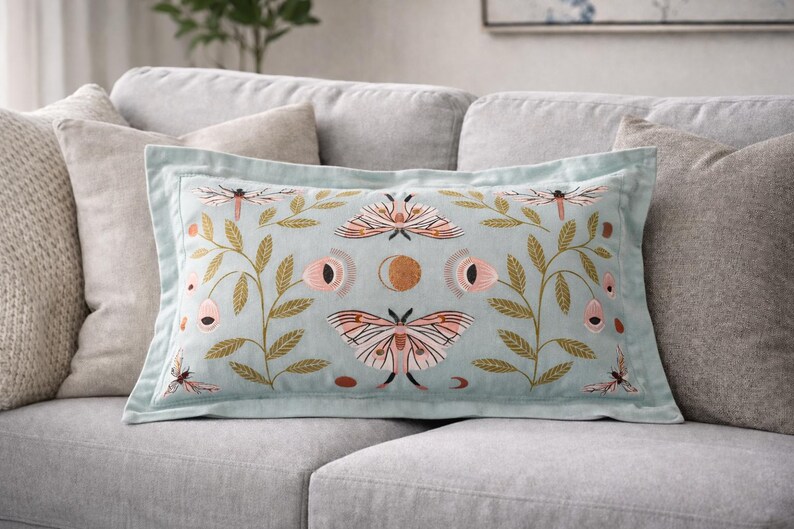 May include: Rectangular cushion with a light blue background and embroidered design. The design features moths, dragonflies, and floral elements in pink, gold, and brown. The cushion has a light blue border and is placed on a grey sofa.