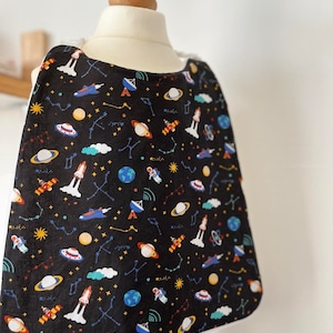 Puede incluir: Un babero negro con ribete blanco que presenta un estampado de temática espacial. El estampado incluye planetas, estrellas, cohetes, astronautas y satélites sobre un fondo negro.