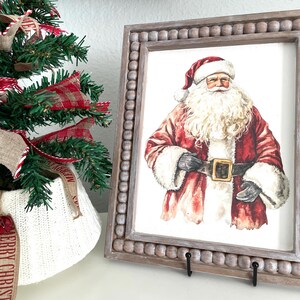 May include: A framed Santa Claus print with a beaded wooden frame. The Santa illustration features a red suit, white beard, and a black belt with a gold buckle. A small Christmas tree with red and white ribbon is to the left.