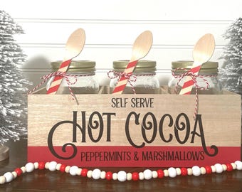 May include: A wooden hot cocoa station with three glass jars, each with a wooden spoon and red and white twine. The wooden box has the words "Self Serve Hot Cocoa" in black and red. Two small, silver Christmas trees flank the box.