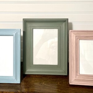 May include: Three rectangular picture frames in varying sizes and colors. The frames are painted in light blue, olive green, and dusty rose. Each frame has a white interior, ready for a photo or artwork.