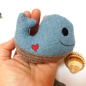 May include: A handcrafted blue whale soft toy with a red heart. The whale features a brown base and a smiling face with black eyes. A seashell is next to the whale.