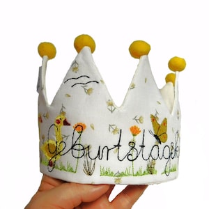 Puede incluir: Una corona de tela blanca con pompones de fieltro amarillo y flores bordadas, un pájaro, una mariposa y la palabra "Geburtstag".