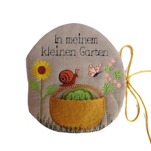 Könnte beinhalten: Besticktes Leinen-Gartenschild mit dem Text "In meinem kleinen Garten". Mit einer Sonnenblume, einer Schnecke, einem Schmetterling und einem Korb mit grünem Gemüse. Das Schild hat eine gelbe Schnur zum Aufhängen.