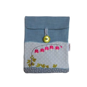 Puede incluir: Una bolsa de tela azul con un patrón de lunares blancos y un diseño de flores bordadas en rosa y verde. La bolsa tiene un cierre de botón y una pequeña etiqueta que dice "Nellie Sue".