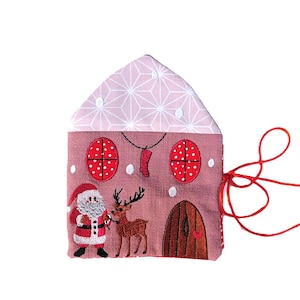 Könnte beinhalten: Ein rosa-roter, weihnachtlicher Stoffschmuck in Form eines Hauses mit einem Weihnachtsmann- und Rentiermotiv. Der Schmuck hat eine rote Schnur.