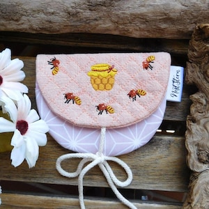 May include: A small, quilted, light pink pouch featuring an embroidered yellow honey pot and several bees on the flap. The pouch has a white geometric pattern on the lower section, a white drawstring closure, and a white tag labeled NestStern.