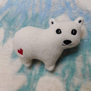 May include: A small, white, plush polar bear toy with a soft, textured surface. The bear features two black button eyes, a stitched black nose and mouth, and a small, red embroidered heart on its rear hip area, facing toward the right side.