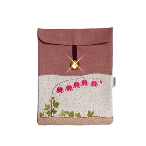 May include: A pink linen envelope with a gold button closure and a delicate embroidered design of pink bleeding hearts on a beige background. The envelope has a small white tag with the text "Needle & Thread".