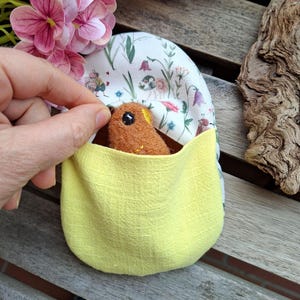 May include: A small, brown felt bird emerges from a yellow pouch. The pouch is lined with a floral fabric with small flowers and insects. The bird has a black eye and a yellow beak. The pouch is held open by a hand.
