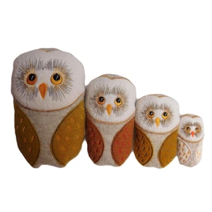 Puede incluir: Un conjunto de cuatro peluches con forma de búho de diferentes tamaños. Los búhos tienen cabezas blancas con detalles bordados, ojos grandes amarillos y picos naranjas. Sus cuerpos son una mezcla de beige, naranja y marrón amarillento.