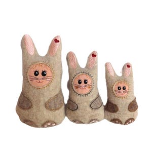 Puede incluir: Tres juguetes de peluche de conejo de diferentes tamaños, todos hechos de fieltro beige con orejas rosas y caras bordadas. El conejo más grande tiene un corazón rojo en su oreja.