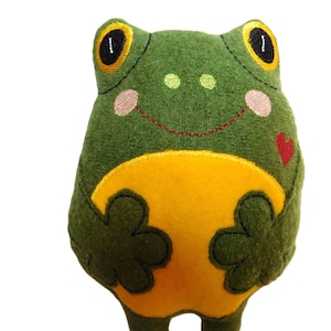 May include: A handmade plush frog toy in shades of green and yellow. The frog has large, expressive eyes with black pupils and yellow irises. It features a smiling mouth, pink cheeks, and a small red heart. The frog's belly is yellow.