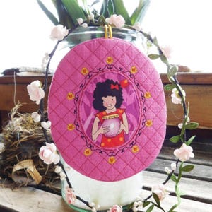 Pode incluir: Um protetor de panela oval rosa acolchoado com uma ilustração de desenho animado de uma menina segurando uma bola de cristal. O protetor de panela tem uma alça amarela para pendurar e é decorado com motivos florais amarelos e laranja. O fundo é decorado com flores.