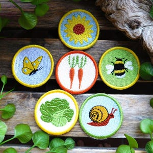 Könnte beinhalten: Eine Sammlung von sechs runden gestickten Aufnähern mit Naturmotiven, darunter eine gelbe Sonnenblume, ein gelber Schmetterling, zwei orangefarbene Karotten, eine schwarz-gelbe Hummel, ein grüner Kopfsalat und eine braune Schnecke.