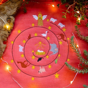 May include: Embroidered artwork on a coral-colored fabric. The design features a spiral with an angel, sheep, and other animals, along with a star and a baby in a manger. Yellow stars are scattered throughout the design.