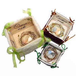 May include: Three fabric Easter baskets with green and brown ribbon ties. The baskets are decorated with embroidered nests and eggs. The largest basket is white with the text "Easter nest" embroidered on it. The medium basket is burgundy with the text "Easter nest" embroidered on it. The smallest basket is white with a black and white nest and eggs embroidered on it.