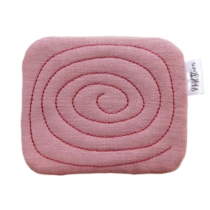 May include: A square, pink fabric coaster with a spiral stitched design in red thread. The coaster has rounded corners and a small white tag with the text "Nest Stern".