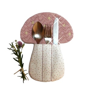 Könnte beinhalten: Ein rosa-weißer Stoff-Besteckhalter in Form eines Pilzes mit einem Tupfenmuster. Eine Gabel, ein Messer und ein Löffel befinden sich im Halter.