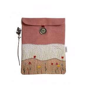 Peut inclure: Une pochette en lin rose de style enveloppe avec une fermeture à bouton. La pochette présente un motif brodé d'un champ de fleurs et d'herbe.