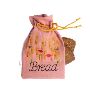 Può includere: Un sacchetto per il pane in lino rosa con un cordoncino di chiusura giallo e spighe di grano e papaveri rossi ricamati. Il sacchetto riporta la scritta "Bread" in corsivo nero.