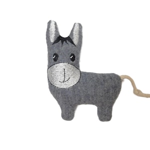 May include: A grey stuffed donkey toy with a silver embroidered face and black eyes. The donkey has a small, beige tail and is made of soft fabric. The toy is designed to stand upright.
