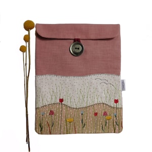 May include: A pink linen laptop sleeve with a button closure and an embroidered floral design. The design features a field of green grass with red, yellow, and blue flowers.