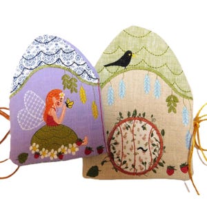 Puede incluir: Dos teteras bordadas. Una lavanda con una hada y una mariposa, la otra beige con un pájaro negro y un diseño floral. Ambas tienen costuras decorativas y detalles de fresas. Las teteras están conectadas con una cuerda amarilla.