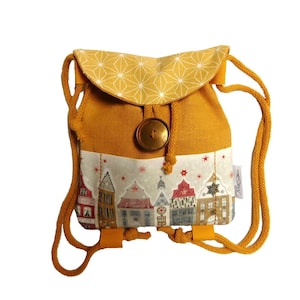 May include: A small yellow drawstring backpack with a white fabric panel featuring a winter village scene. The backpack has a gold button closure and a geometric pattern on the top flap. The text "Nolé Design" is printed on the bottom right corner of the fabric panel.