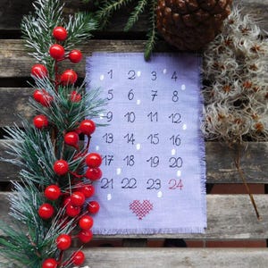 Könnte beinhalten: Ein Adventskalender aus lavendelfarbenem Stoff mit gestickten Zahlen und einem roten Herz. Der Kalender ist mit weißen Punkten und einem roten Herz verziert. Der Hintergrund umfasst einen Kiefernzweig mit roten Beeren, einen Tannenzapfen und getrocknete Blumen.