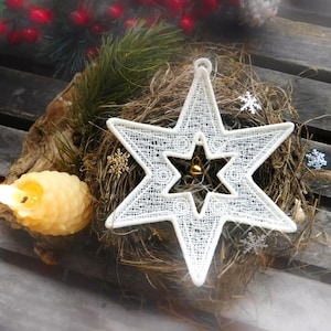 Könnte beinhalten: Ein weißer, sternförmiger Weihnachtsschmuck mit filigranen Spitzendetails. Der Schmuck liegt auf einem Bett aus getrocknetem Gras, mit einer kleinen goldenen Glocke in der Mitte. Eine brennende Kerze und dekorative Elemente ergänzen die festliche Szene.