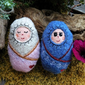 Op de afbeelding: Twee handgemaakte vilten babypoppen. Eén pop is gewikkeld in een roze deken met een hart en de andere in een blauwe deken. Beide poppen hebben geborduurde gezichten en dragen capuchons.