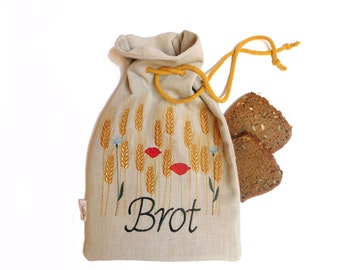 May include: A beige linen bread bag with a yellow drawstring and embroidered wheat stalks, red poppies, and the word "Brot" in black. Two slices of dark bread are next to the bag.