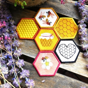 Könnte beinhalten: Eine Sammlung von sechs sechseckigen bestickten Aufnähern, die in einem Wabenmuster angeordnet sind. Die Designs umfassen gelbe Wabenstrukturen, Bienen auf weißem Grund, einen Honigtopf, ein schwarzes Herz auf Weiß und eine Biene auf einer Blume.