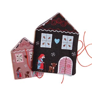 May include: Two felt gingerbread house ornaments with embroidered details. The larger house is brown with a blue heart and snowflakes. The smaller house is pink with a red heart and snowflakes. Both houses have a red string attached.