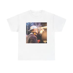 Puede incluir: Una camiseta blanca con una foto de una persona que lleva una gorra de béisbol blanca y un pañuelo, soplando humo de su boca.