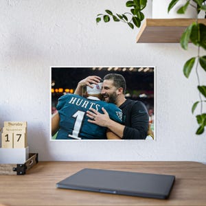 以下が含まれることがあります： 緑と白のユニフォームを着た2人のアメリカンフットボール選手がフィールドで抱き合っています。手前の選手は「HURTS」という名前と背番号「1」のジャージを着ています。