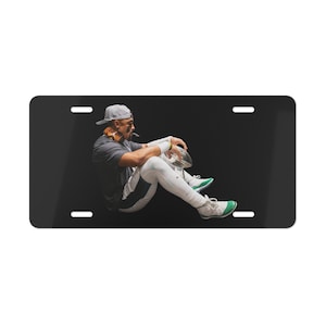May include: A black and white license plate featuring a photo of a person wearing a white baseball cap and a green and white jersey sitting with a football in their hands.