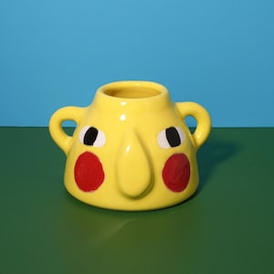 Puede incluir: Una maceta amarilla de cerámica con forma de cara y dos asas. La maceta presenta ojos negros y blancos, una nariz larga y círculos rojos en las mejillas. El fondo es azul y verde contrastantes.