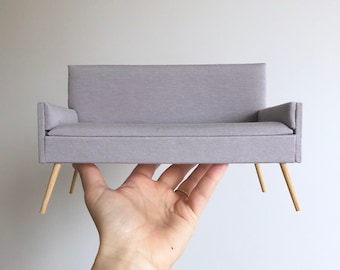 May include: A small, light gray fabric sofa with wooden legs. The sofa is being held in a hand.