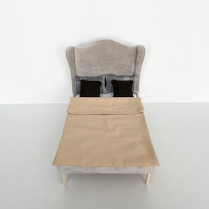 Könnte beinhalten: Ein graues, gepolstertes Bett mit einem geflügelten Kopfteil, einer beigefarbenen Bettdecke und zwei Kissen mit schwarz-weißem Muster.