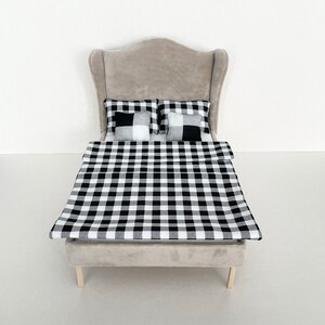 Könnte beinhalten: Ein Puppenbett mit einem grauen Kopfteil und einer schwarz-weiß karierten Bettdecke und Kissen.