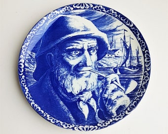Op de afbeelding: Blauw en wit keramisch bord met een gedetailleerde illustratie van een man met een baard en een hoed, die een pijp rookt. De man kijkt naar rechts. De achtergrond toont een schip en een stad.