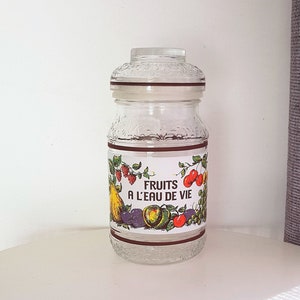 May include: A clear glass jar with a brown lid and a white label that says "Fruits a l'eau de vie". The label has a colorful illustration of various fruits, including cherries, grapes, pears, and plums.