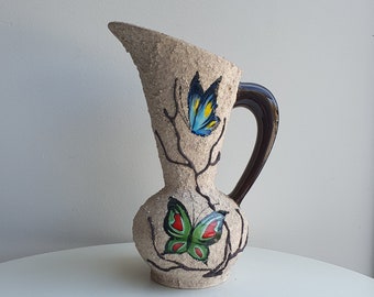 May include: A ceramic pitcher with a brown handle and a textured surface. It is decorated with two painted butterflies, one blue and one green, on a branch design.